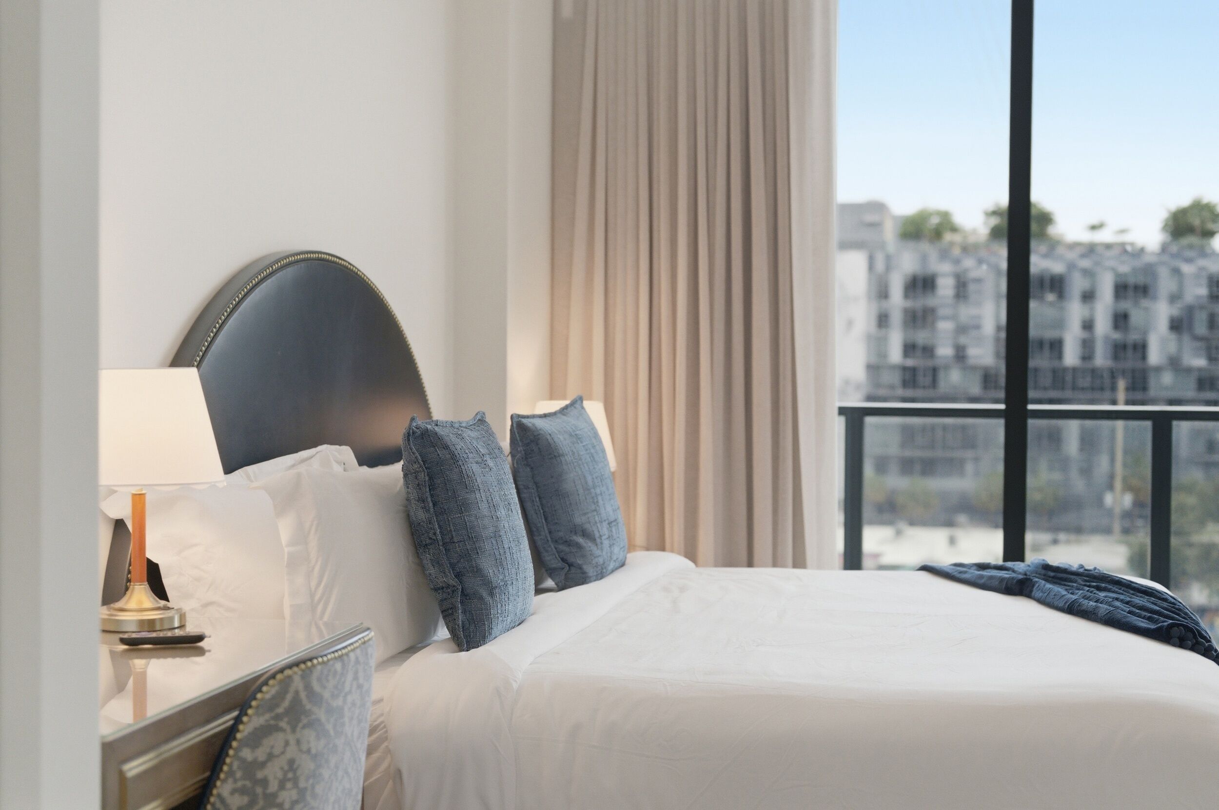 Een hotelkamer met een bed, nachtkastje, lamp en uitzicht op gebouwen door een raam.