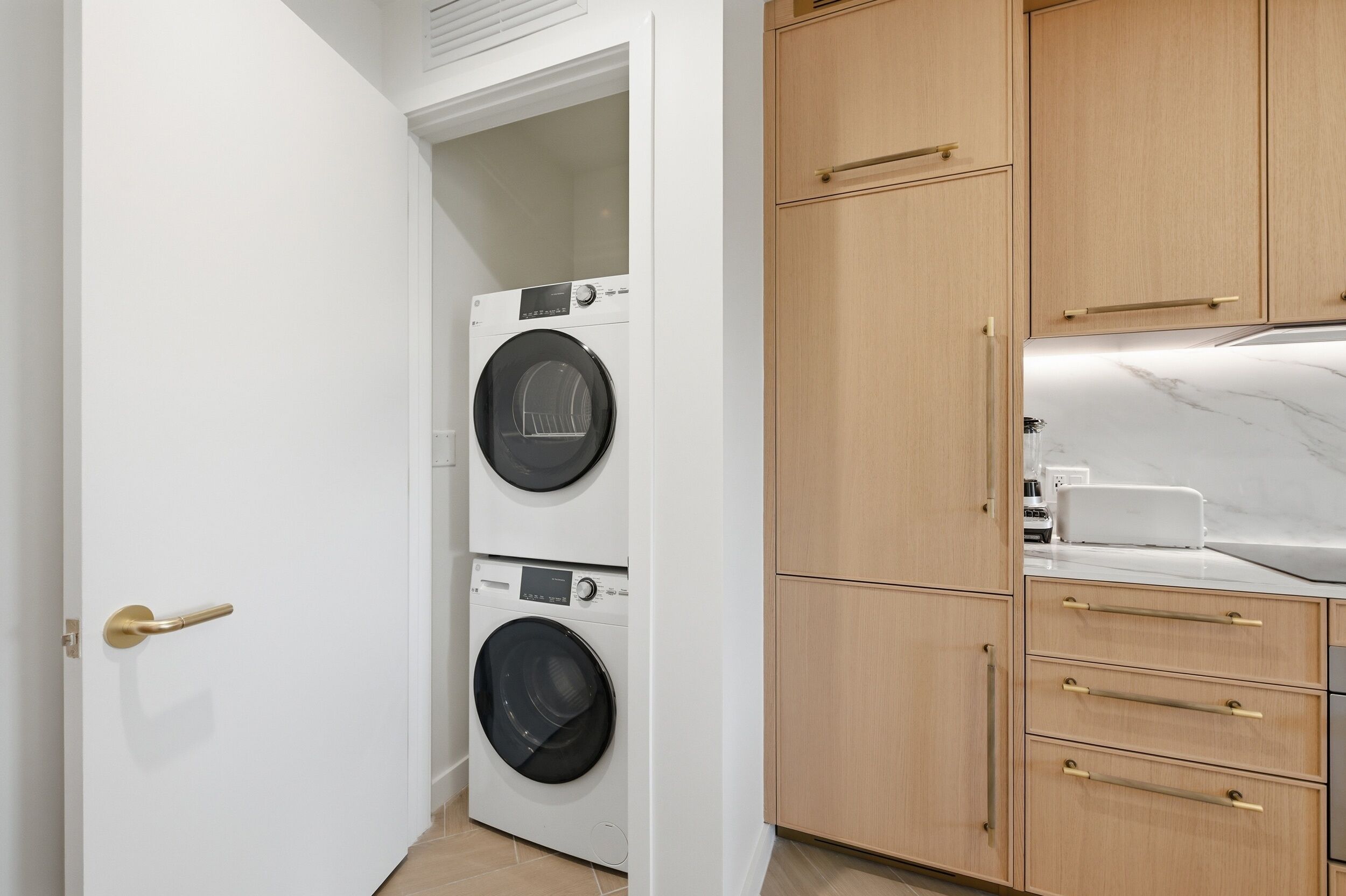 A laundry room with two stacked washing machines, wooden cabinets, and a countertop with a coffee maker.