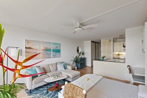 Ein modernes Wohnzimmer mit Sofa, Couchtisch und Bett. Ein Deckenventilator, ein Essbereich mit Stuhl und eine durchgehende Küche sind ebenfalls zu sehen.