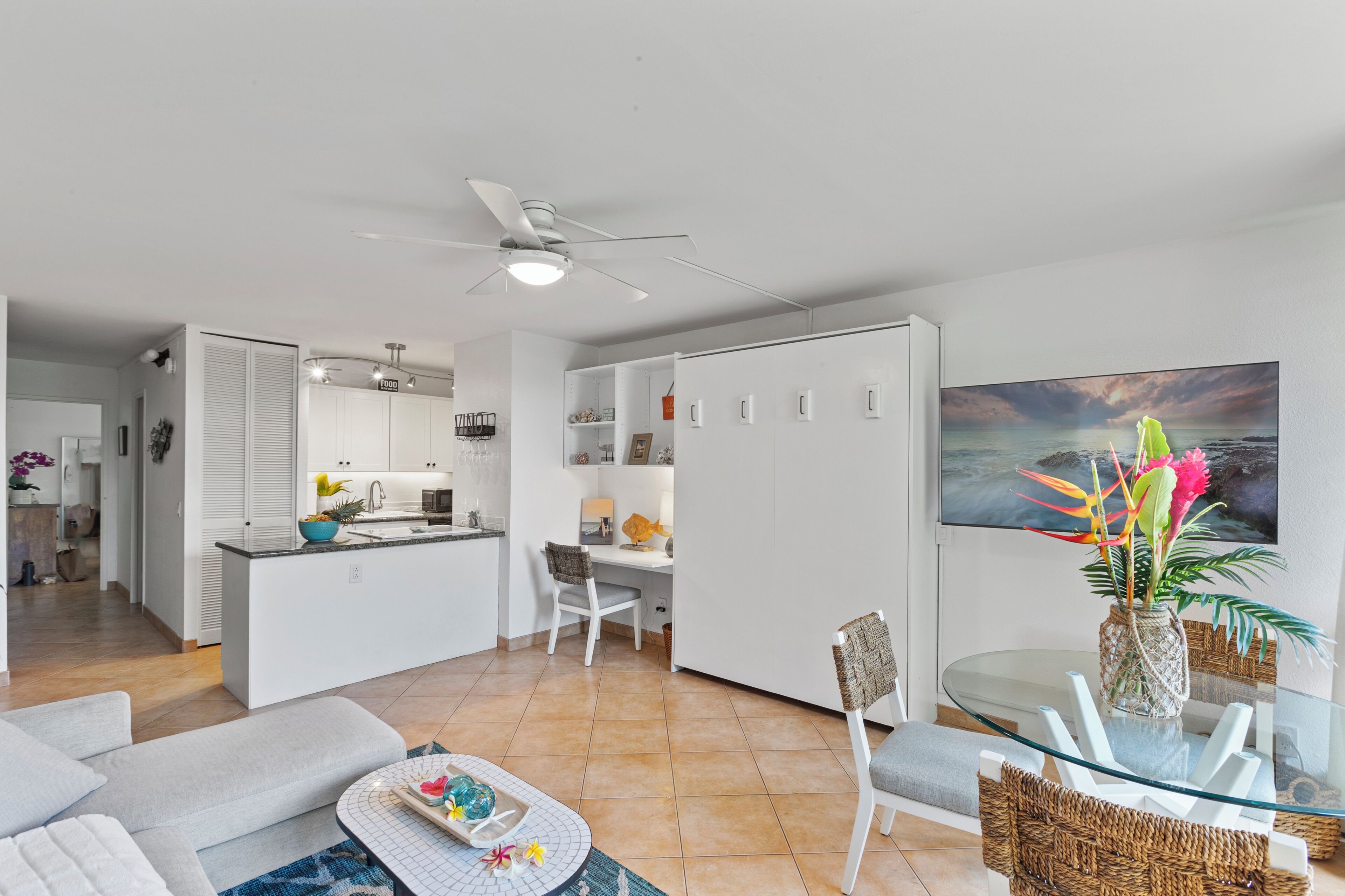 A modern kitchen with white cabinets, a dining area with a glass table, a sofa, and a ceiling fan.
