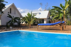 A pool area with a wooden deck, a lounge chair, and a white building with a triangular roof.