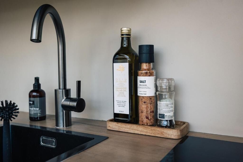 Una encimera de cocina con un grifo moderno, una botella de aceite de oliva, un salero y un pequeño bote de jabón de manos.