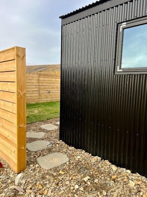 A corrugated metal building with a window, wooden fence, and stone stepping stones. - Night & Day Lodge (Borgarbyggð)