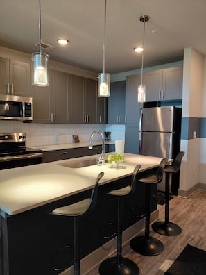 A modern kitchen with stainless steel appliances, a central island with bar stools, pendant lighting, and a tiled backsplash.