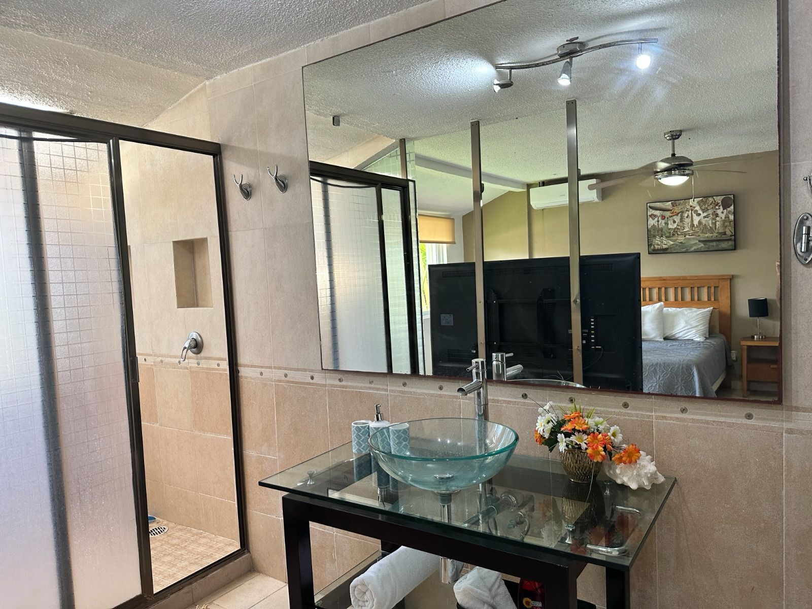A bathroom with a glass bowl sink, a mirror, a shower, and a bed with a wooden headboard.
