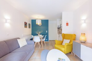 Ein Wohnzimmer mit einer Couch, einem gelben Sessel, einem Esstisch mit Stühlen und einer Küchenzeile im Hintergrund.