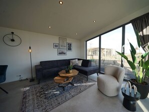 Een moderne woonkamer met een bank, salontafel en stoel, grote ramen en uitzicht op de omgeving.