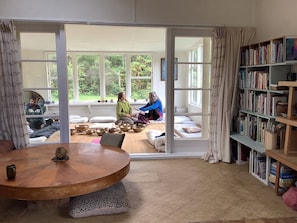 Un intérieur comprenant une table ronde en bois, une bibliothèque et, à travers de grandes portes vitrées, une vue sur un espace extérieur aménagé avec des coussins et des bols.