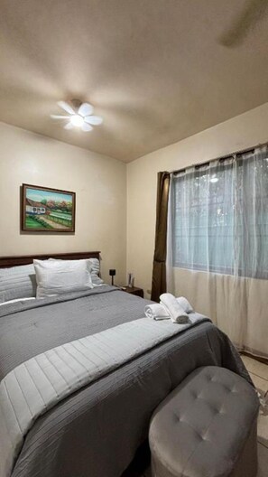 Une chambre à coucher comprenant un lit, une table de chevet, une lampe, une fenêtre avec des rideaux et un ventilateur de plafond.