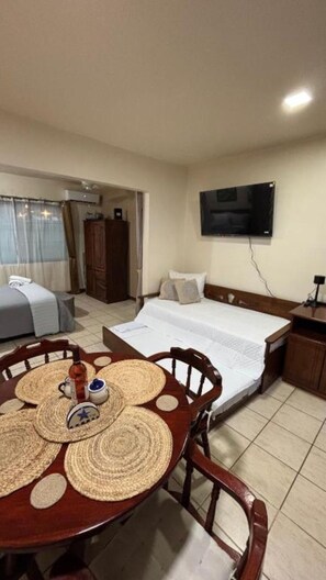 Habitación de hotel con cama, televisor montado en la pared, una mesa de comedor con individuales y una ventana con cortinas.