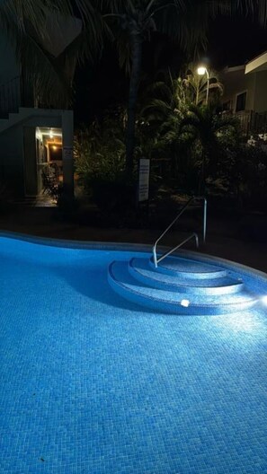 Una piscina con escalones de acceso, rodeada de palmeras y un edificio con una entrada iluminada.