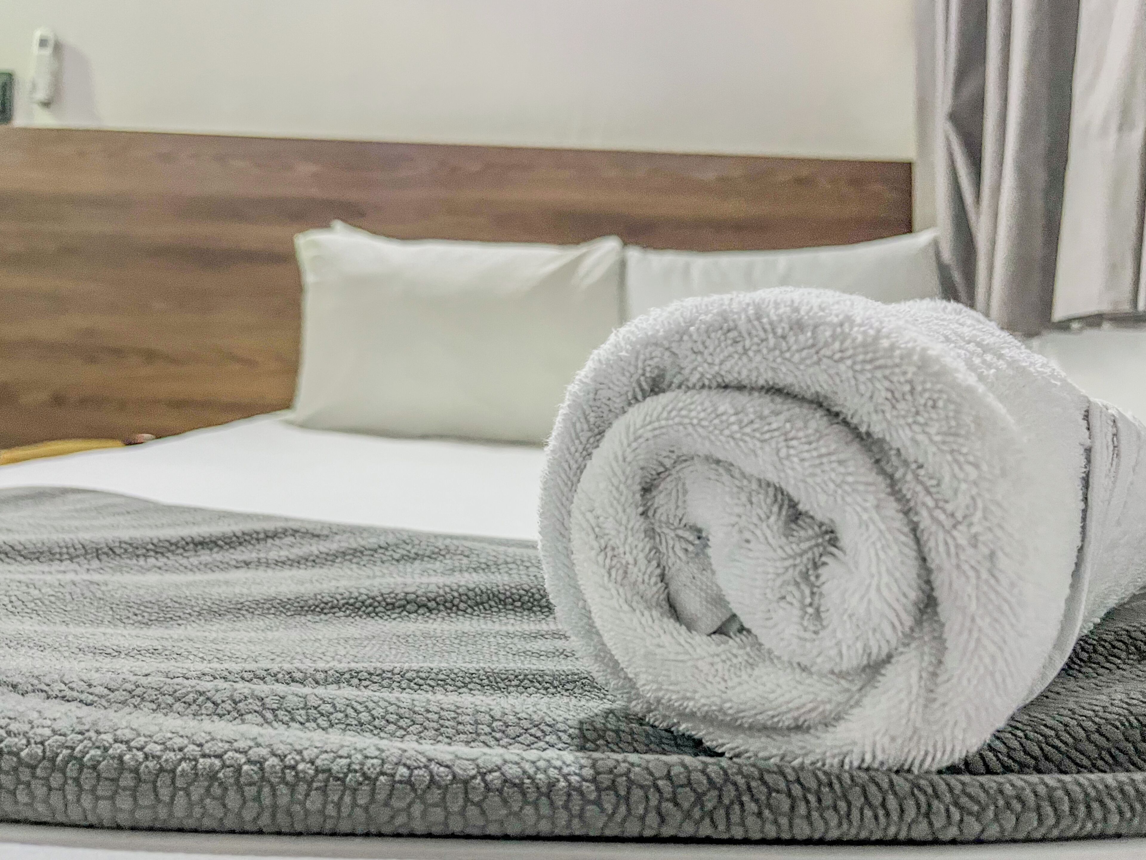 A neatly made bed with a rolled white towel, a grey blanket, and a wooden headboard.