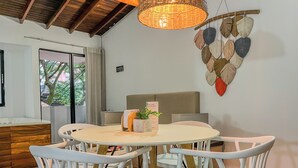 Un coin repas avec une table ronde, des chaises blanches, un luminaire en fibres tressées et une décoration murale représentant des feuilles.