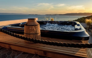 A hot tub with a view of the sea and hills, surrounded by a wooden deck and a rope barrier.