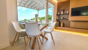Un comedor moderno con una mesa de vidrio y sillas blancas, un televisor montado en la pared y vistas al exterior.