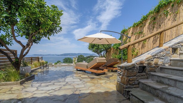 Un patio de piedra con sillones de descanso y una sombrilla, con vistas a un cuerpo de agua y un paisaje montañoso.