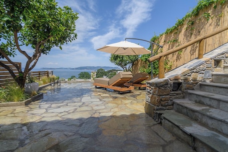 A stone patio with lounge chairs and an umbrella, overlooking a body of water and a mountainous landscape.. LemonTree Villa Glossa Skopelos