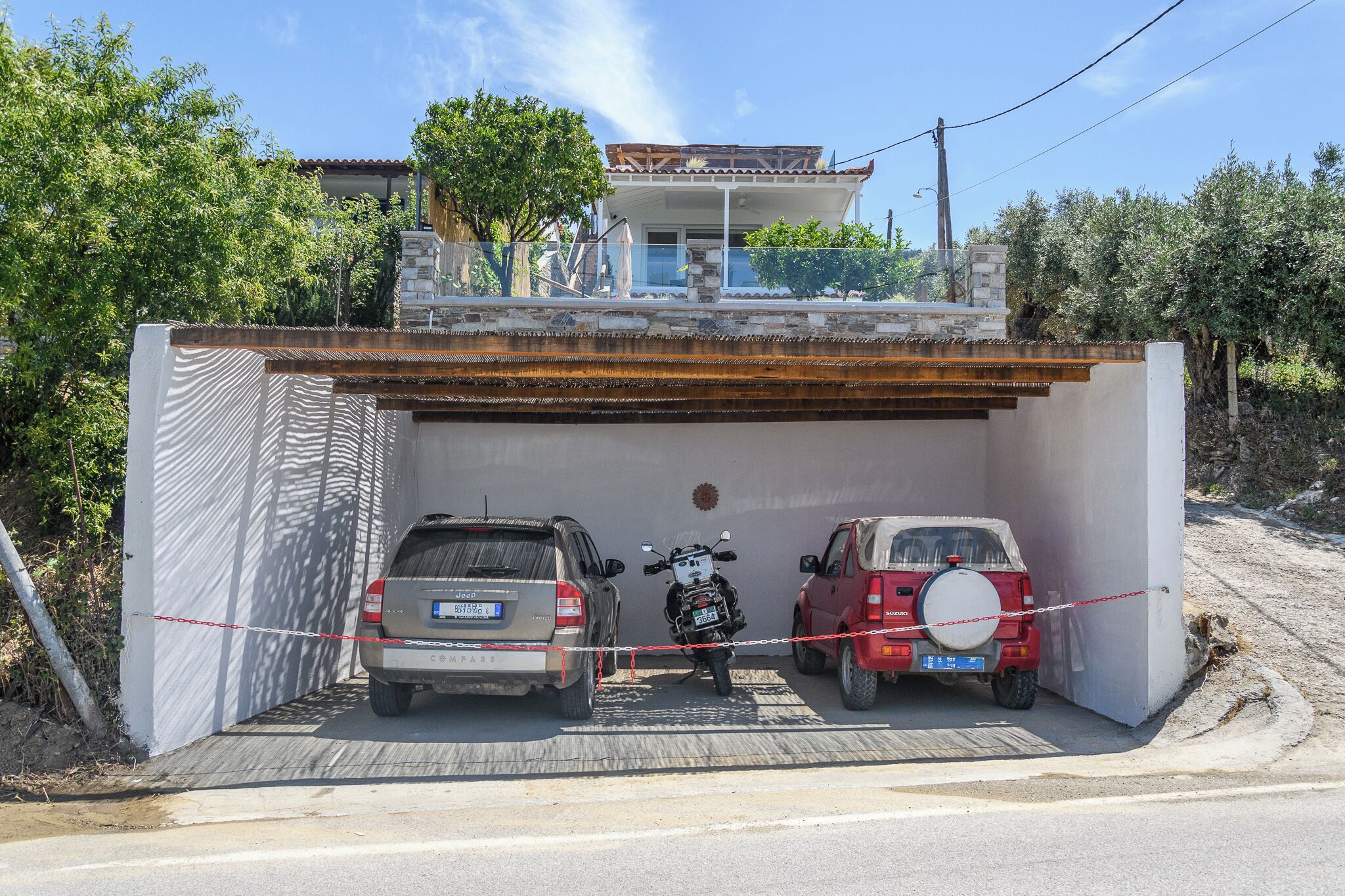 Un garage abritant deux voitures et une moto, une maison en arrière-plan et un ciel dégagé.
