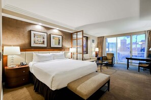Ein Hotelzimmer mit einem großen Bett, einem Schreibtisch, einem Sessel, einer Couch und Blick auf die Stadt durch große Fenster.