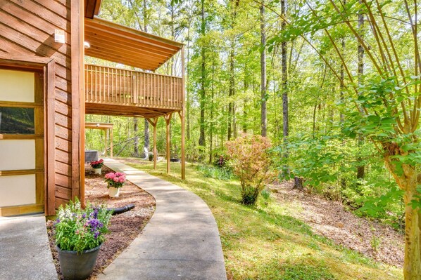 A wooden cabin with a covered porch, a concrete pathway, and a forested area with various plants and trees.