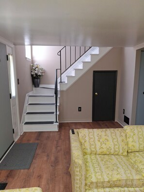 Eine Treppe mit weißen Stufen und schwarzen Geländern führt zu einer dunklen Tür. Im Vordergrund steht ein gelb gemustertes Sofa, und auf der Zwischenebene befindet sich ein Blumentopf.