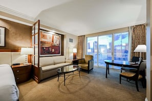 Ein Hotelzimmer mit Sofa, Sessel, Couchtisch, Schreibtisch und einem großen Fenster mit Blick auf die Stadt.
