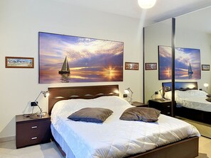 Ett sovrum med en säng, sängbord, lampor och en stor spegel som reflekterar en målning av en solnedgång.