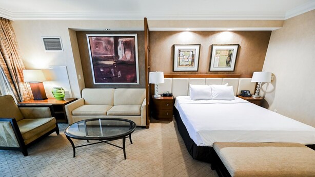 A hotel room with a bed, a sofa, a chair, a small table, and two bedside lamps. - SVS: Strip View Studio | Signature at MGM (Las Vegas)