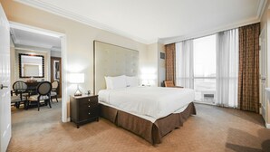 Ein Hotelzimmer mit einem großen Bett, einem Nachttisch mit Lampe, einem Sessel, einer Essecke mit Tisch und Stühlen sowie Blick ins Freie durch das Fenster.