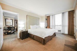 Ein Hotelzimmer mit einem großen Bett, einem Nachttisch mit Lampe, einem Sessel, einer Essecke mit Tisch und Stühlen sowie Blick ins Freie durch das Fenster.