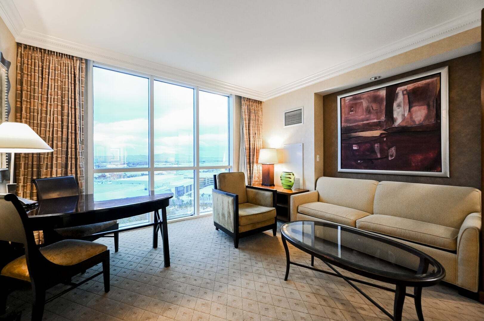 Une chambre d’hôtel avec une grande fenêtre, un bureau, un canapé, une table basse et un tableau au mur.