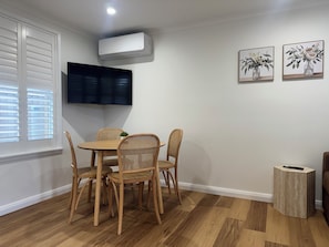 Un comedor con una mesa redonda, cuatro sillas de mimbre, un televisor montado en la pared y dos cuadros de flores también colgados en la pared.