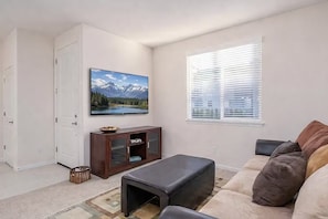 A living room with a sofa, ottoman, TV, cabinet, and a window with blinds. - 2 BR Layout with Bonus Room • Quiet + Parking (Carson City)