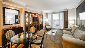 Una camera d'hotel con zona pranzo, divano, televisore e un'ampia finestra con tende.