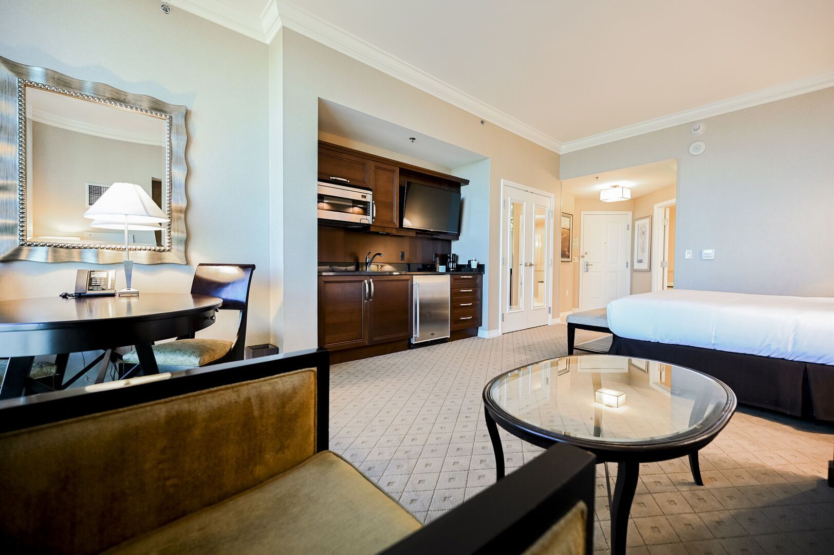 A hotel room with a bed, a round glass table, a sofa, a kitchen area, and a large mirror.