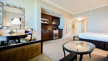 A hotel room with a bed, a round glass table, a sofa, a kitchen area, and a large mirror.