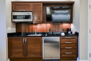 A compact kitchen with a microwave, a built-in TV above the counter, a stainless steel refrigerator, and wooden cabinetry. - SVS: Sphere Strip View Balcony | Signature at MGM (Las Vegas)