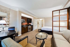 A hotel room with a bed, a sofa, a dining table with chairs, a kitchen area, and a large window with blinds. - SVS: Sphere Strip View Balcony | Signature at MGM (Las Vegas)