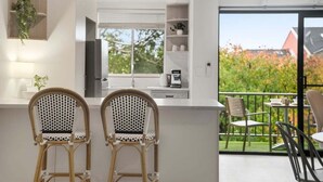 Una cocina moderna con dos sillas de mimbre, un balcón con mobiliario exterior y vistas a árboles y edificios.