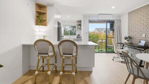 Una cocina moderna con armarios blancos, un comedor con dos sillas de mimbre y vistas a un balcón con pared de ladrillo.