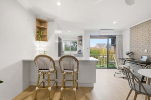 A modern kitchen with white cabinetry, a dining area with two wicker chairs, and a view of a balcony with a brick wall.