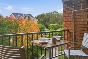 A balcony with a table set for dining, a chair, and a view of trees and buildings.