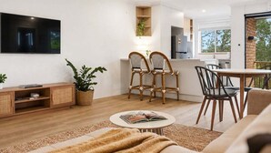 Una sala de estar moderna con un sofá, una mesa de centro con un libro, dos sillones de mimbre, un televisor montado en la pared y una cocina visible a través de un acceso abierto.
