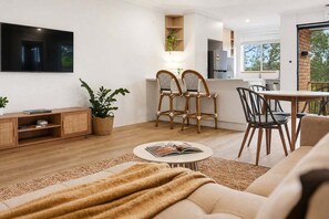 A modern living room with a sofa, a coffee table with a book, two wicker chairs, a TV mounted on the wall, and a kitchen area visible through an open door. - Riviera - Stylish Interiors, Sleeps 4,  Walk to Beach (Coffs Harbour)