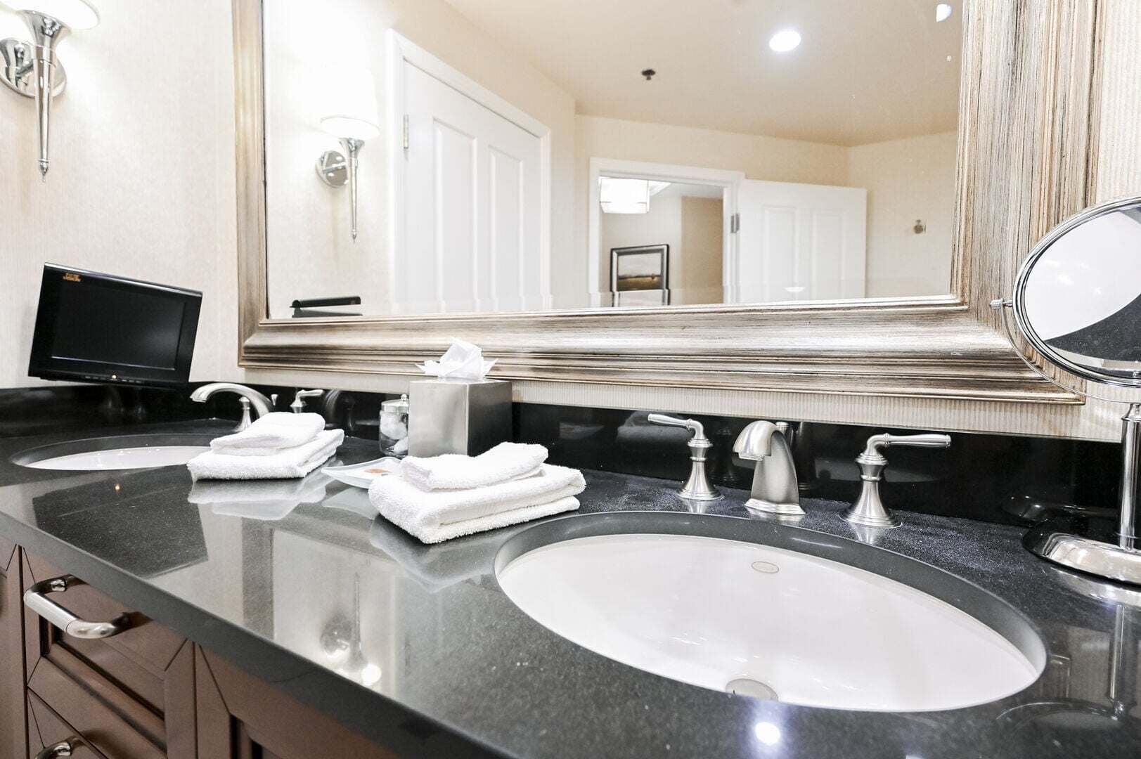 A hotel bathroom with two sinks, a large mirror, and a television.