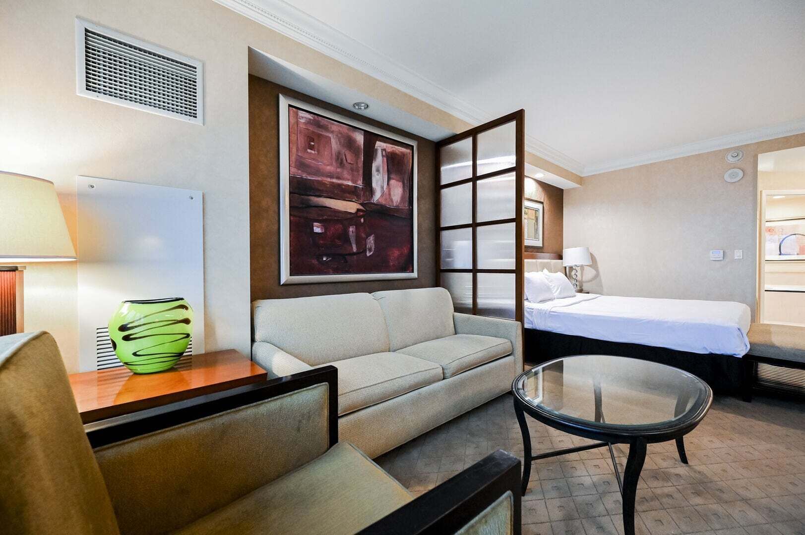A hotel room with a sofa, a glass table, a bed, a painting, and a lamp.