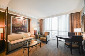 Ein Hotelzimmer mit einer Couch, einem kleinen Tisch, einem Esstisch mit Stühlen, einem großen Fenster mit Vorhängen und einem Bild an der Wand.