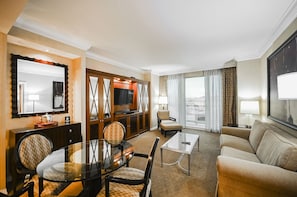 Ein Hotelzimmer mit Essbereich, einer Couch, einem Fernseher und einem großen Fenster mit Vorhängen.