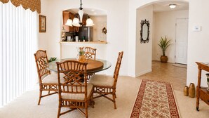 A dining area with a round table and four chairs, a kitchen in the background, a mirror on the wall, and a potted plant.