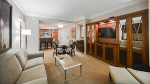 Ein Wohnzimmer mit Sofa, Glastisch, Esstisch mit Stühlen, einer Küchenzeile und einem Fernseher in einem Holzkabinett.
